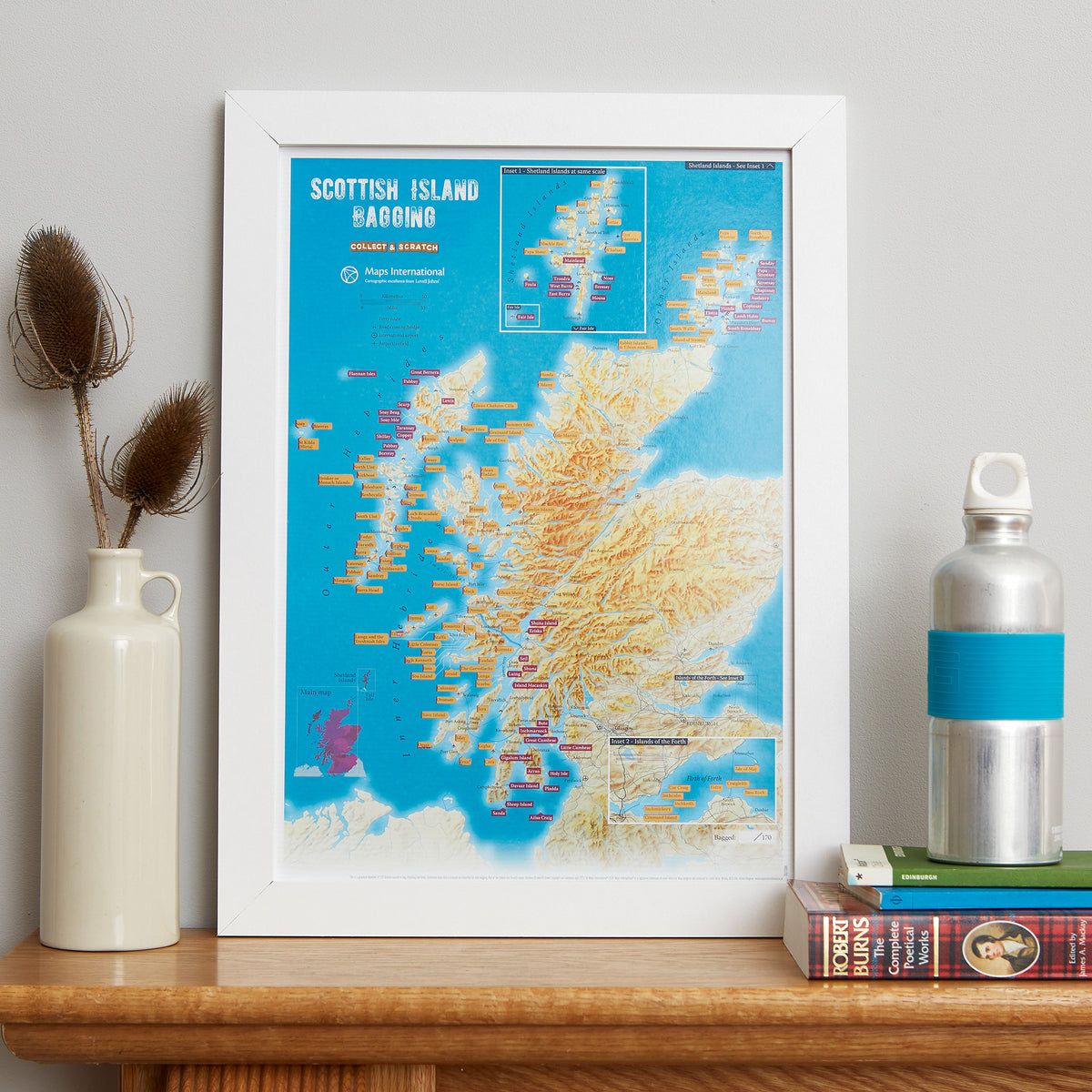 A framed Scottish Island Hiking map print leaning on a wooden shelf, styled with a cream vase holding dried stems on the left and a silver water bottle on books on the right.